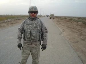 Soldier standing on deserted road, military vehicle behind.