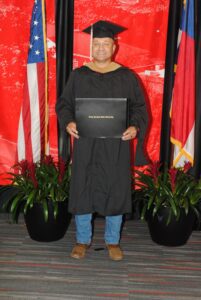 Person holding a diploma in front of American flags and red backdrop.