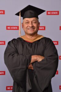 Smiling graduate in cap and gown with arms crossed at NC State.