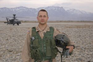 Soldier in military gear holding helmet in a mountainous desert landscape.