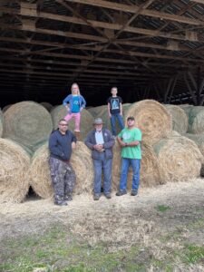 Five men standing in front of large hay bales inside a barn.